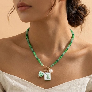 May include: A necklace featuring a strand of small green beads and a gold-toned circular clasp. The pendant arrangement includes a white heart-shaped charm with green floral patterns, a white mahjong tile with green bamboo, and a white pearl.