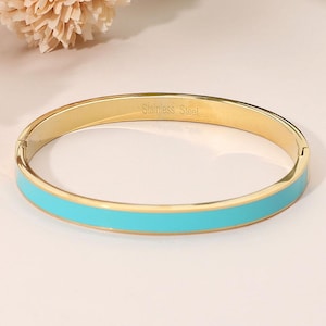May include: A gold-colored stainless steel bangle bracelet with a turquoise enamel band. The words "Stainless Steel" are engraved on the inside of the bracelet. The bracelet is a hinged style.