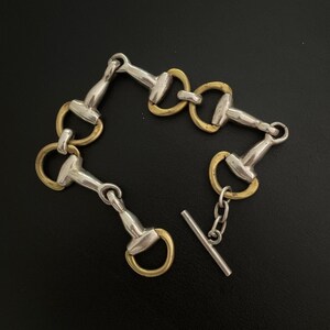 May include: A link bracelet featuring a repeating horse bit design composed of alternating silver-toned metal bars and circular gold-toned rings. The piece includes a toggle clasp closure with a silver-toned bar and a short chain link connector.