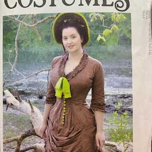 May include: McCall's Costumes pattern featuring a woman in a brown dress with a green bow and buttons. The dress has a ruffled neckline and sleeves. The pattern includes the text "Angela Clayton".