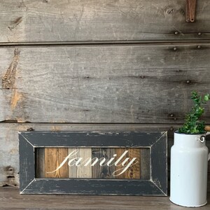 May include: A rectangular wooden sign with the word "family" in white script. The sign is framed in a distressed dark gray, with a wooden background of various wood tones. A white enamel pitcher with a wooden handle and greenery sits to the right.