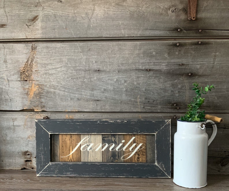 May include: A rectangular wooden sign with the word "family" in white script. The sign is framed in a distressed dark gray, with a wooden background of various wood tones. A white enamel pitcher with a wooden handle and greenery sits to the right.