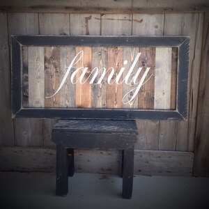 May include: A rustic wooden sign with the word "family" in elegant white script. The sign features a black frame and is made of vertical wooden planks in various natural tones. It is displayed on a small black wooden stool.
