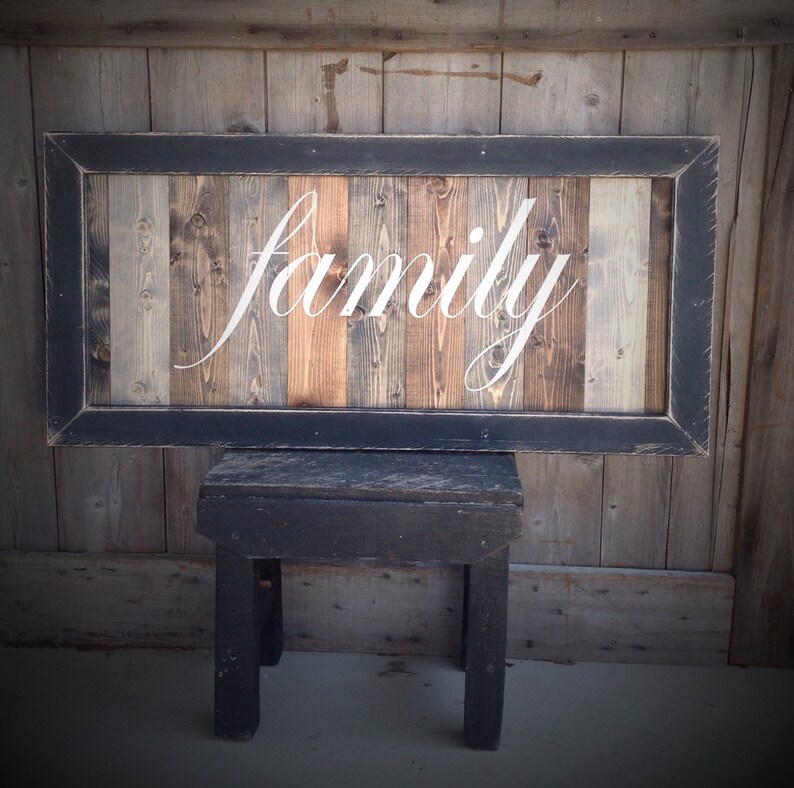 May include: A rustic wooden sign with the word "family" in elegant white script. The sign features a black frame and is made of vertical wooden planks in various natural tones. It is displayed on a small black wooden stool.