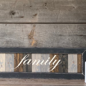 May include: A rectangular wooden sign with the word "family" in white script. The sign has a black frame and a background of vertical wooden planks in shades of gray and brown. A white milk jug with greenery sits to the right.