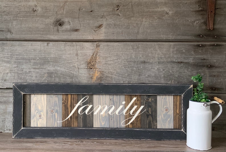 May include: A rectangular wooden sign with the word "family" in white script. The sign has a black frame and a background of vertical wooden planks in shades of gray and brown. A white milk jug with greenery sits to the right.