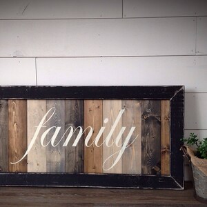 May include: A rectangular wooden sign with the word "family" in white script. The sign has a black frame and a background of vertical wooden planks in shades of brown, gray, and beige. A small metal bucket with greenery sits to the right.