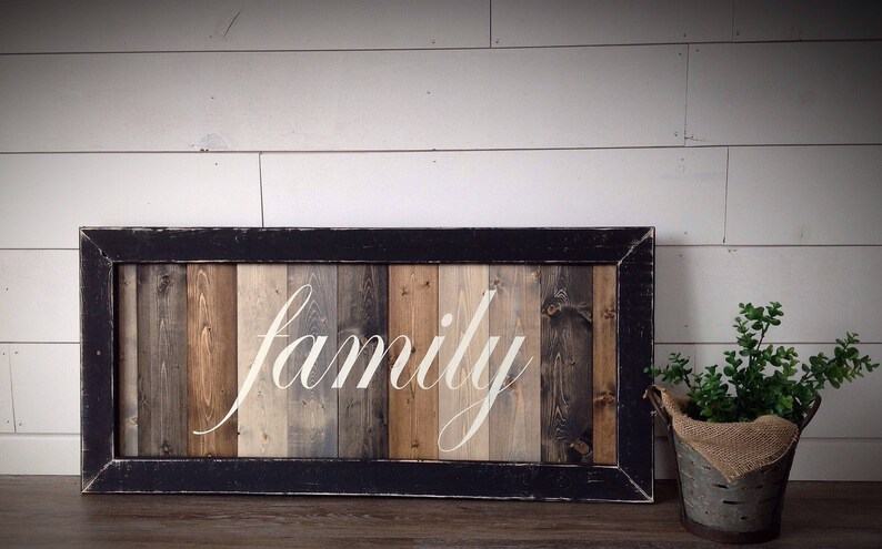 May include: A rectangular wooden sign with the word "family" in white script. The sign has a black frame and a background of vertical wooden planks in shades of brown, gray, and beige. A small metal bucket with greenery sits to the right.