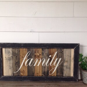 May include: A rectangular wooden sign with the word "family" in white script. The sign is framed in black and features vertical wooden planks in various shades of brown and gray. A small potted plant sits to the right.