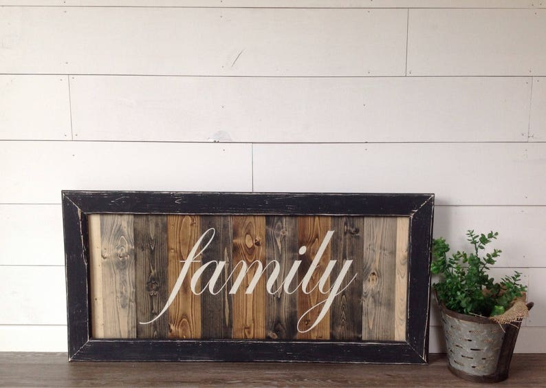 May include: A rectangular wooden sign with the word "family" in white script. The sign is framed in black and features vertical wooden planks in various shades of brown and gray. A small potted plant sits to the right.