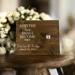 May include: A rustic wooden sign with the text "AND THE two SHALL BECOME one" and the names "Francisco & Joselyn" with the date "March 14, 2026" in white script. A small metal clasp is attached to the right side of the sign.