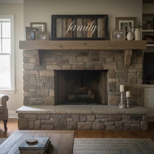 May include: A stone fireplace with a wooden mantel and a framed sign that says "family". The fireplace has a large opening and is flanked by a window and a built-in cabinet with a television. There are framed photos and candles on the mantel.