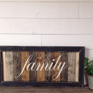 May include: Wooden framed sign with the word "family" in white script. The sign has a black frame and a background of vertical wooden planks in various shades of brown and gray. A small metal bucket with greenery is to the right.