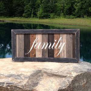 May include: A rustic wooden sign with the word "family" in white script. The sign is framed in black and features vertical wooden planks in various shades of brown and gray. The sign is displayed on a large rock.