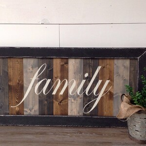 May include: A rectangular wooden sign with the word "family" in white script. The sign has a black frame and is made of vertical wooden planks in shades of brown and gray. A metal bucket with greenery sits to the right.
