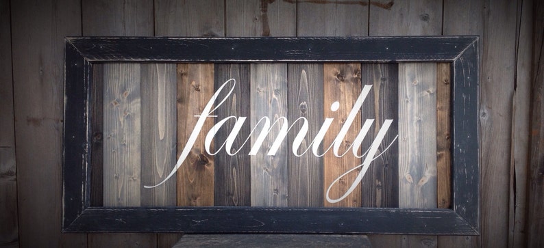 May include: Wooden sign with the word "family" in white script. The sign has a black frame and is made of vertical wooden planks in shades of brown, gray, and natural wood tones. The background is a wooden wall.