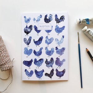 May include: A white notebook with a blue and white illustration of roosters on the cover. The notebook says "NOTES" on the front. The notebook is surrounded by craft supplies, including a spool of twine, two tubes of paint, a paintbrush, and a blue pencil.