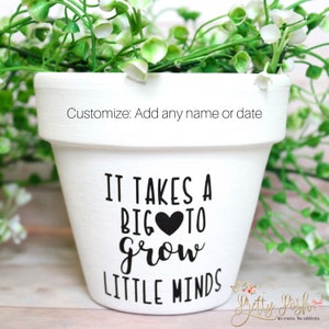 May include: White ceramic flower pot with black text that reads "It takes a big heart to grow little minds". The pot is decorated with a black heart.