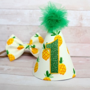 May include: A white and yellow pineapple-patterned party hat with a green tulle pom-pom on top. The hat has a green glitter number "1" on the front. A matching bow tie is also included.