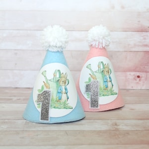 Może przedstawiać: Dwa niebieskie i różowe filcowe czapki imprezowe z białym pomponem na górze. Każda czapka ma motyw z Peterem Królikiem z numerem jeden z srebrnego brokatu.