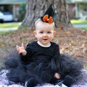 Puede incluir: Un beb&eacute; vestido con un disfraz de bruja negro, que incluye un vestido negro, un tut&uacute; y un sombrero puntiagudo con un lazo naranja. El beb&eacute; est&aacute; sentado en una manta floral al aire libre.