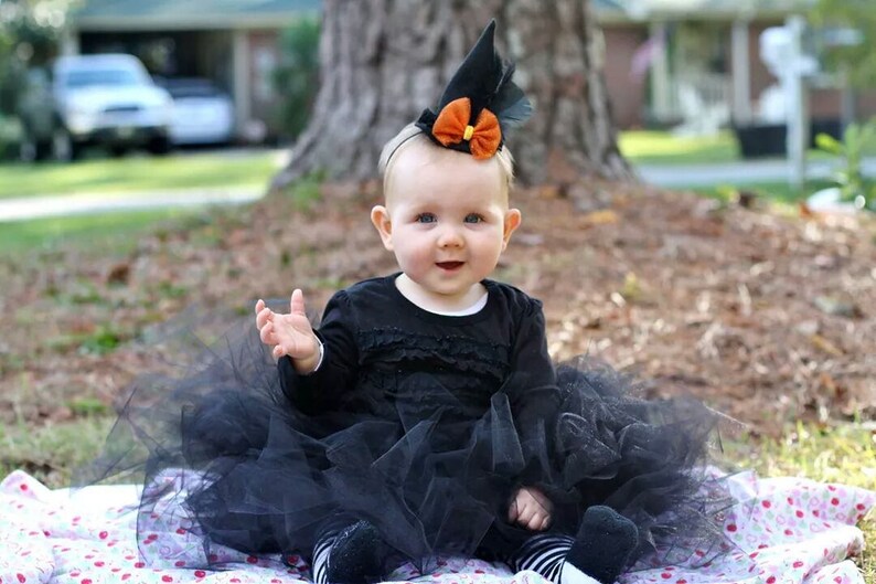 Puede incluir: Un beb&eacute; vestido con un disfraz de bruja negro, que incluye un vestido negro, un tut&uacute; y un sombrero puntiagudo con un lazo naranja. El beb&eacute; est&aacute; sentado en una manta floral al aire libre.