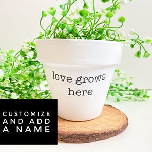 May include: White ceramic flower pot with the text "love grows here" on the front. The pot is filled with a green plant and is sitting on a wooden coaster. The text "CUSTOMIZE AND ADD A NAME" is visible in the lower left corner of the image.
