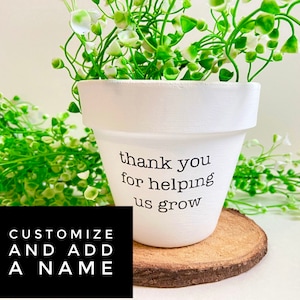 May include: White ceramic planter with the text "thank you for helping us grow". The planter is filled with a small green plant with white flowers and is sitting on a wooden coaster.