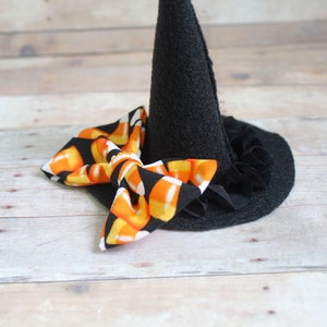Puede incluir: Un peque&ntilde;o sombrero de bruja negro con un lazo de caramelos de ma&iacute;z. El sombrero tiene una corona c&oacute;nica y un ala ancha con un borde con volantes. El lazo presenta un patr&oacute;n de caramelos de ma&iacute;z en naranja, amarillo y blanco.