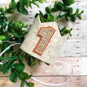 Może przedstawiać: Korona z filcu z numerem jeden z juty. Korona jest biała z pomarańczowymi szwami wokół numeru. Korona jest związana białą wstążką.