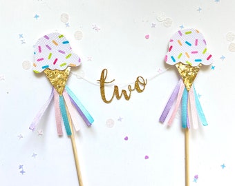 Könnte beinhalten: Zwei Eiswaffel-Kuchentopper mit Streuseln und goldenen Glitzerkegeln. Die Kegel sind an Holzspießen mit pastellrosa, blauen und violetten Bändern befestigt. Das Wort "zwei" ist in goldenen Glitzer zwischen den Kegeln geschrieben.