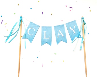Op de afbeelding: Een blauwe en witte taarttopper met het woord "CLAY" gespeld op een banner. De banner is bevestigd aan twee houten stokjes met blauwe en witte linten.