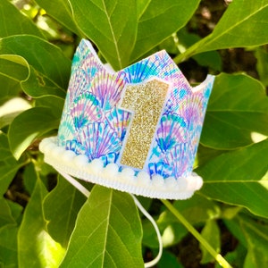 May include: A white crown with a blue and purple seashell pattern and a gold glitter number one on top. The crown has a white pom-pom trim.