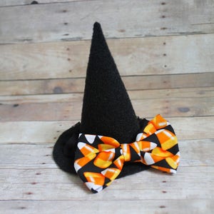 Puede incluir: Un sombrero de bruja negro con un lazo de caramelos de ma&iacute;z. El sombrero tiene una corona en forma de cono y un ala ancha. El lazo es negro con un patr&oacute;n de caramelos de ma&iacute;z naranja, amarillo y blanco.