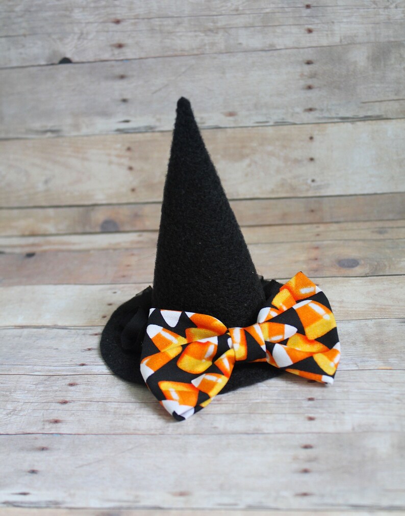 Puede incluir: Un sombrero de bruja negro con un lazo de caramelos de ma&iacute;z. El sombrero tiene una corona en forma de cono y un ala ancha. El lazo es negro con un patr&oacute;n de caramelos de ma&iacute;z naranja, amarillo y blanco.