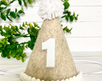 Pode incluir: Um chapéu de festa de feltro bege em forma de cone com um pompom branco no topo e um número "1" de feltro branco na frente. O chapéu tem uma faixa elástica branca e uma borda de pompom branco ao redor da parte inferior.