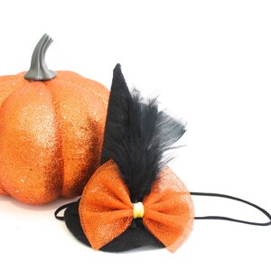 Puede incluir: Imagen con temática de Halloween con un pequeño sombrero de bruja negro con un lazo naranja y pluma negra, junto a una calabaza naranja brillante. El sombrero tiene una banda elástica negra. La calabaza es un objeto decorativo.