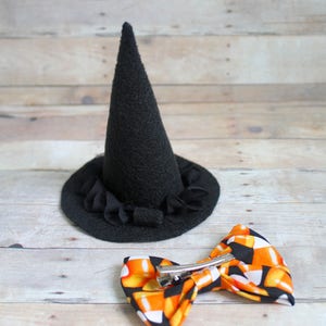 Puede incluir: Un sombrero de bruja de fieltro negro con un borde con volantes y un lazo con estampado de caramelos de ma&iacute;z con un clip de metal. Accesorios tem&aacute;ticos de Halloween sobre un fondo de madera clara.