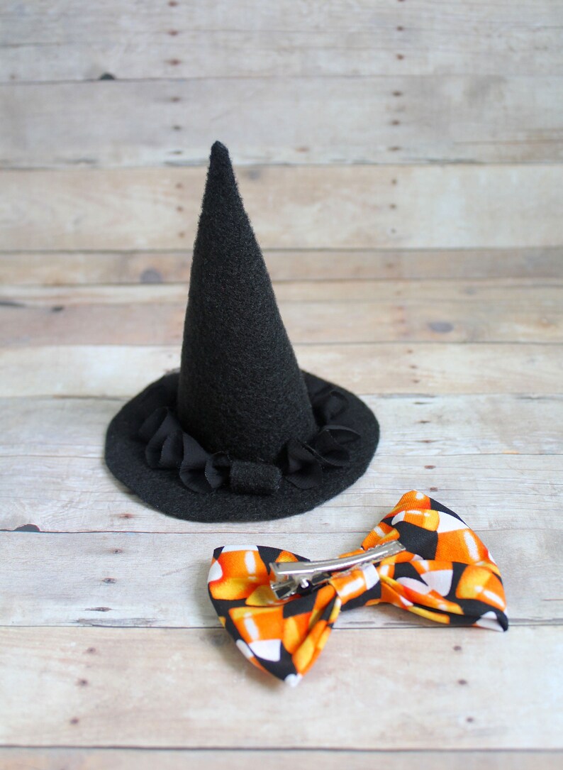 Puede incluir: Un sombrero de bruja de fieltro negro con un borde con volantes y un lazo con estampado de caramelos de ma&iacute;z con un clip de metal. Accesorios tem&aacute;ticos de Halloween sobre un fondo de madera clara.