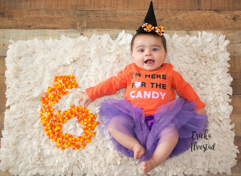 Puede incluir: Un beb&eacute; con una camiseta naranja con el texto "I'M HERE FOR THE CANDY" y un tut&uacute; morado, con un sombrero de bruja negro con un lazo naranja. Un n&uacute;mero "6" hecho de caramelos de ma&iacute;z est&aacute; al lado del beb&eacute;.