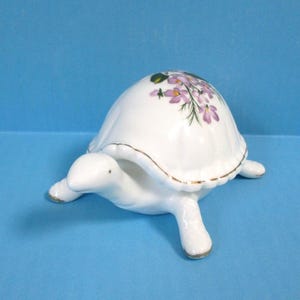 May include: A white ceramic turtle figurine with a floral design on its shell. The turtle has a gold trim and is set against a blue background. The figurine is a decorative item.