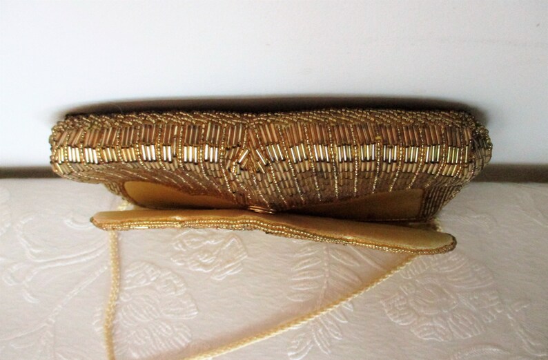 May include: A gold beaded clutch purse with a rectangular shape and a long, thin strap. The purse is covered in small, shiny gold beads, giving it a textured appearance. The purse is likely vintage.