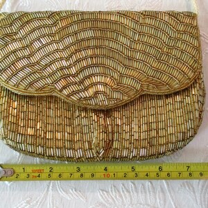May include: A gold beaded clutch purse with a flap closure and a cream-colored shoulder strap. The purse is approximately 8 inches wide. The beads are arranged in vertical and wave-like patterns, creating a textured surface. The purse is on a white surface.