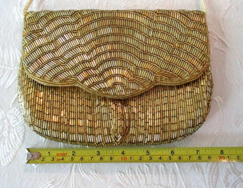 May include: A gold beaded clutch purse with a flap closure and a cream-colored shoulder strap. The purse is approximately 8 inches wide. The beads are arranged in vertical and wave-like patterns, creating a textured surface. The purse is on a white surface.