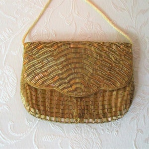 May include: A gold beaded clutch purse with a flap closure and a cream-colored cord strap. The purse is covered in small, vertical beads, creating a textured, wave-like pattern. The overall design is elegant and vintage.
