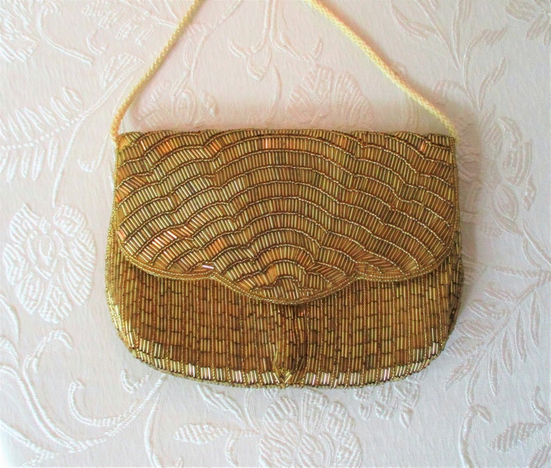 May include: A gold beaded clutch purse with a flap closure and a cream-colored cord strap. The purse is covered in small, vertical beads, creating a textured, wave-like pattern. The overall design is elegant and vintage.