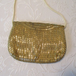 May include: A gold beaded handbag with a cream-colored shoulder strap. The bag is covered in vertical gold beads, creating a textured surface. The bag's shape is rounded at the bottom and has a curved top.