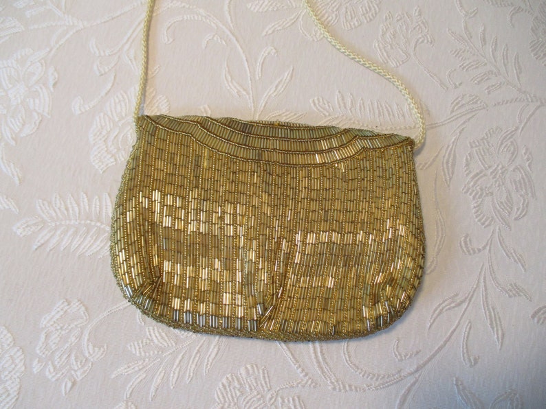 May include: A gold beaded handbag with a cream-colored shoulder strap. The bag is covered in vertical gold beads, creating a textured surface. The bag's shape is rounded at the bottom and has a curved top.