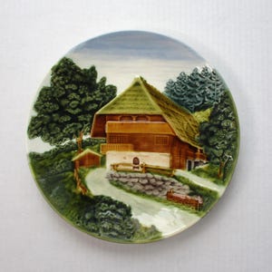 May include: A ceramic plate featuring a 3D image of a brown and green cottage with a thatched roof, surrounded by trees and a stone wall. The plate has a white background with a blue sky.