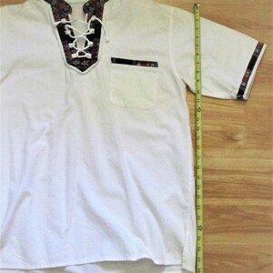 May include: White short-sleeved shirt with a lace-up neckline and a patterned trim. The shirt has a chest pocket with a patterned trim. The shirt is 28 inches long.